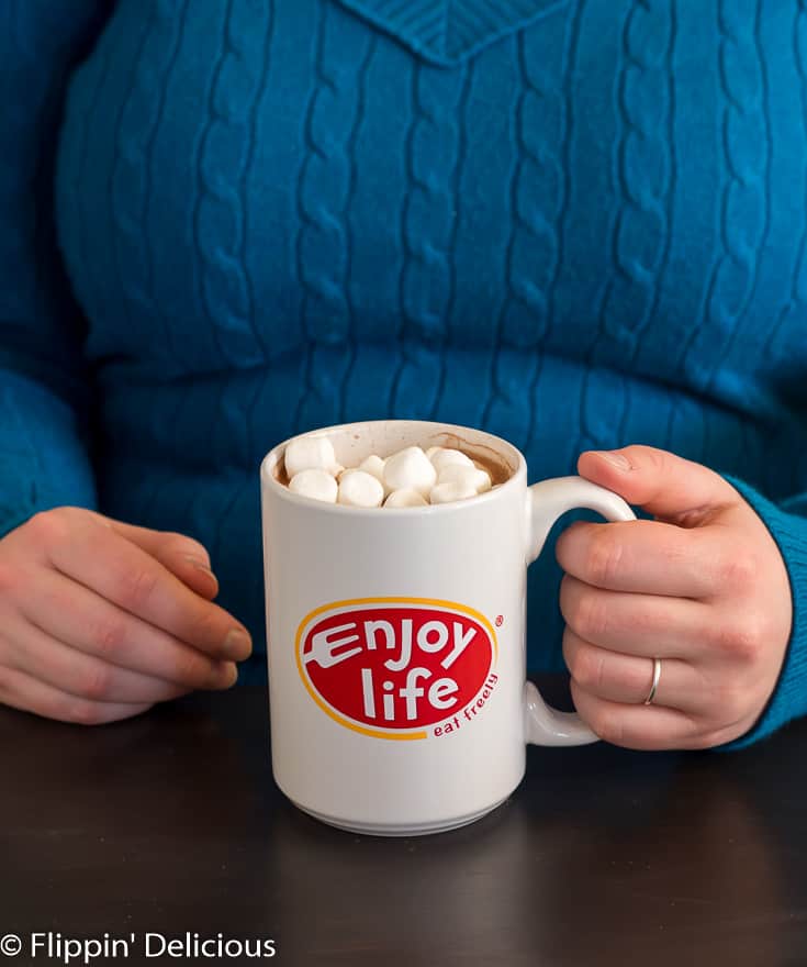 person wearing teal sweater holding white mug with Enjoy Life logo filled with vegan hot chocolate topped with marshmallows
