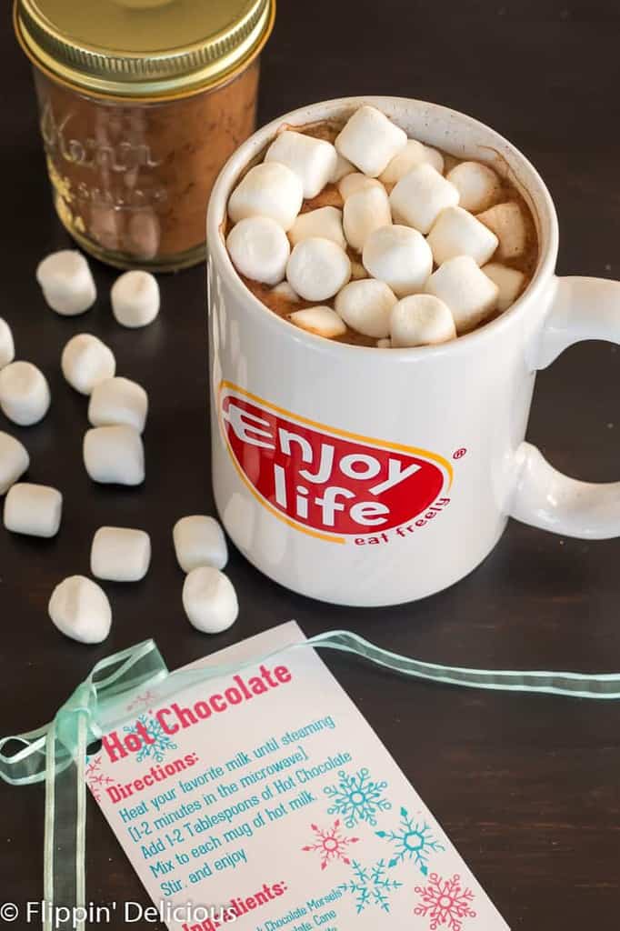 mug of vegan hot chocolate topped with marshmallows on brown table with jar of vegan hot chocolate mix and vegan hot chocolate mix label 