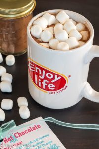 mug of vegan hot chocolate topped with marshmallows on brown table with jar of vegan hot chocolate mix and vegan hot chocolate mix label