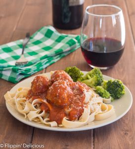 a tan plate with gluten free taglietelle topped with gluten free turkey meatballs and tomatoe sauce with some broccoli, on a brown wooden table with a green checked napkin and a glass of red win in the background