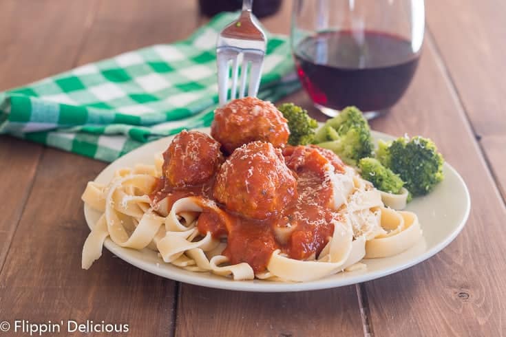 fork reaching for gluten free  meatballs on a bed of gluten free fettucine with tomato sauce with a green and white checked napkin and glass of red wine in the background