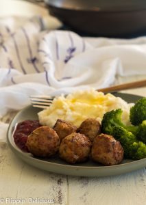 green plate with five sauteed turkey meatballs with a side of red berry sauce, steamed broccoli, and mashed potatoes with butter with a fork and white napkin with purple flowers