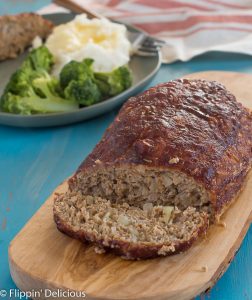 gluten free turkey meatloaf with slice cut and laying beside the ground turkey meatloaf sitting on a wooden cutting board on top of a turquoise table with a green plate with turkey meatloaf, mashed potatoes, and broccoli in the background