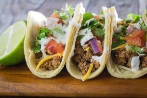 corn tortilla tacos filled with slow cooker ground turkey taco meat, cheese, sour cream, lettuce, tomato, and red onion on a wooden cutting board