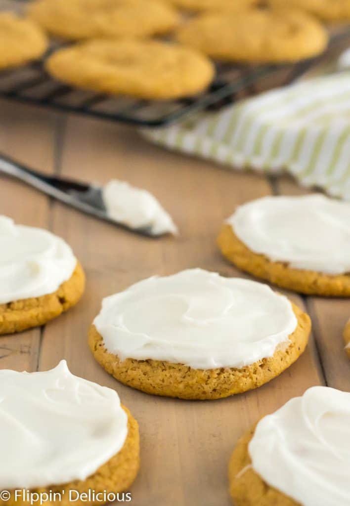 vegan gluten free pumpkin cookies with frosting on a wooden table with cooling rack full of un-frosted cookies and knife with frosting in the background