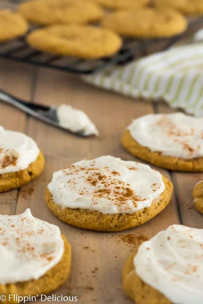 vegan gluten free pumpkin cookies with frosting and a sprinkle of nutmeg on a wooden table with cooling rack full of un-frosted cookies and knife with frosting in the background