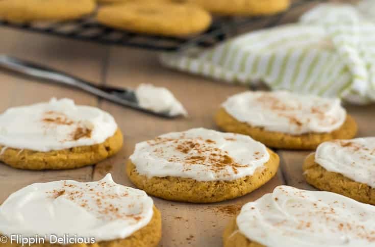 vegan gluten free pumpkin cookies with frosting and a sprinkle of nutmeg on a wooden table with cooling rack full of un-frosted cookies and knife with frosting in the background