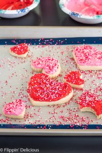 pink and red gluten free heart sugar cookies on a silicone baking sheet on a cookie sheet with pink, white, and red non pareil sprinkles