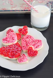 gluten free heart sugar cookies with pink and red frosting and pink, white, and red non pareil sprinkles on a white plate with a scalloped edge on a dark wooden table with a glass of milk with a white and red striped straw in the background