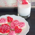 gluten free heart sugar cookies with pink and red frosting and pink, white, and red non pareil sprinkles on a white plate with a scalloped edge on a dark wooden table with a glass of milk with a hand dipping a red heart cookie with a bite taken out of it, with a white and red striped straw in the background