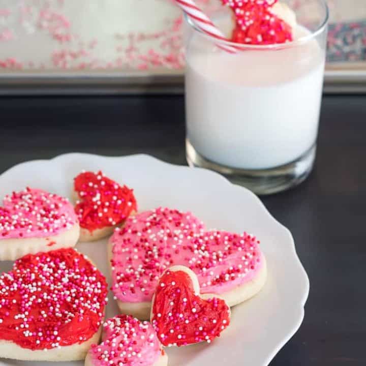 gluten free heart sugar cookies with pink and red frosting and pink, white, and red non pareil sprinkles on a white plate with a scalloped edge on a dark wooden table with a glass of milk with a hand dipping a red heart cookie with a bite taken out of it, with a white and red striped straw in the background