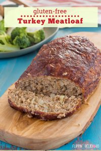 gluten free turkey meatloaf with a slice cut off, beside the meatloaf, on a wooden cutting board with a green plate with broccoli, mashed potatoes and gluten free meatloaf in the background, on a teal wooden table with gray streaks with text "gluten-free turkey meatloaf"