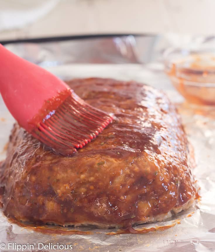 ground turkey meatloaf on a baking sheet lined with aluminum foil being brushed with glaze using a red silicone basting brush with small bowl of glaze in the background