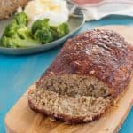 gluten free turkey meatloaf with a slice cut off, beside the meatloaf, on a wooden cutting board with a green plate with broccoli, mashed potatoes and gluten free meatloaf in the background, on a teal wooden table with gray streaks
