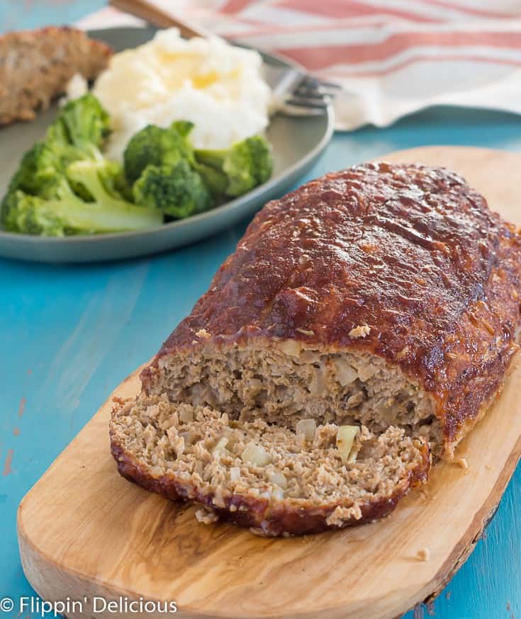 gluten free turkey meatloaf with a slice cut off, beside the meatloaf, on a wooden cutting board with a green plate with broccoli, mashed potatoes and gluten free meatloaf in the background, on a teal wooden table with gray streaks