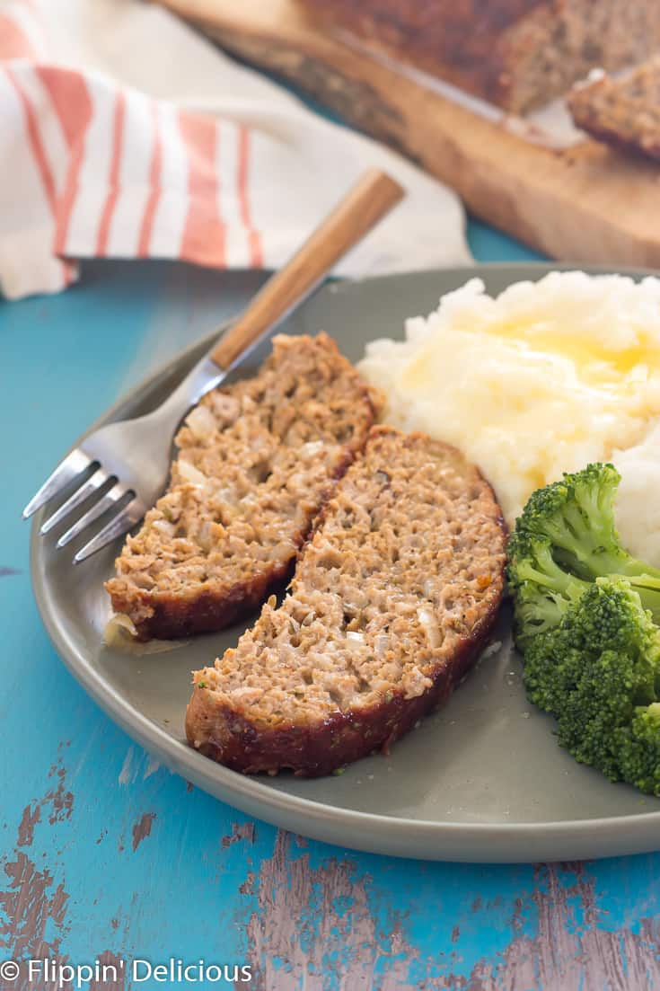 two slices of gluten free turkey meatloaf on a green plate with mashed potatoes and broccoli and a wooden-handled fork on a teal table with gray streaks with a white napkin with an orange stripe in the background