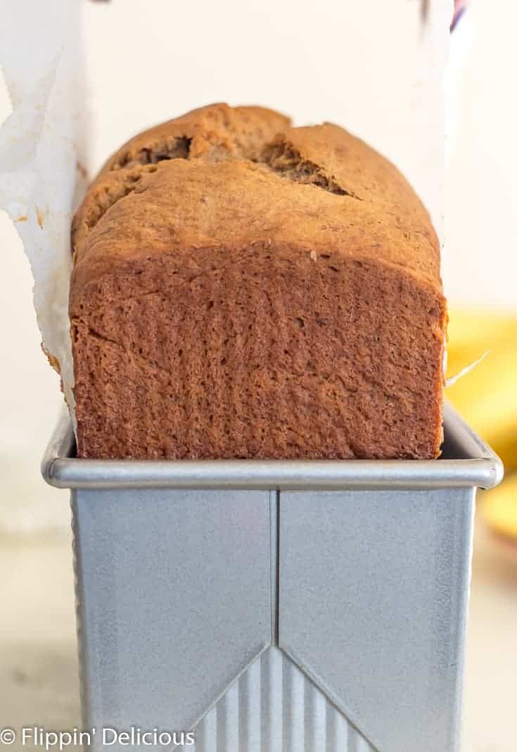 lifting gluten free banana bread from metal 9x4 loaf pan using parchment paper with a bunch of bananas in the background