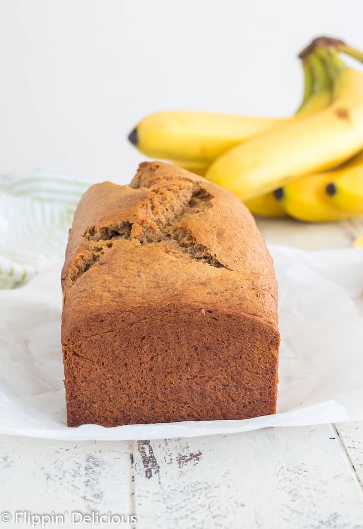 un-sliced loaf of gluten free banana bread on a piece of parchment paper on a white wooden table with a bunch of ripe bananas in the background