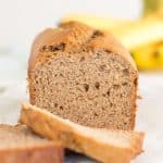loaf of gluten free banana bread with a few slices in front and out of focus with a bunch of bananas in the background, on a white wooden table