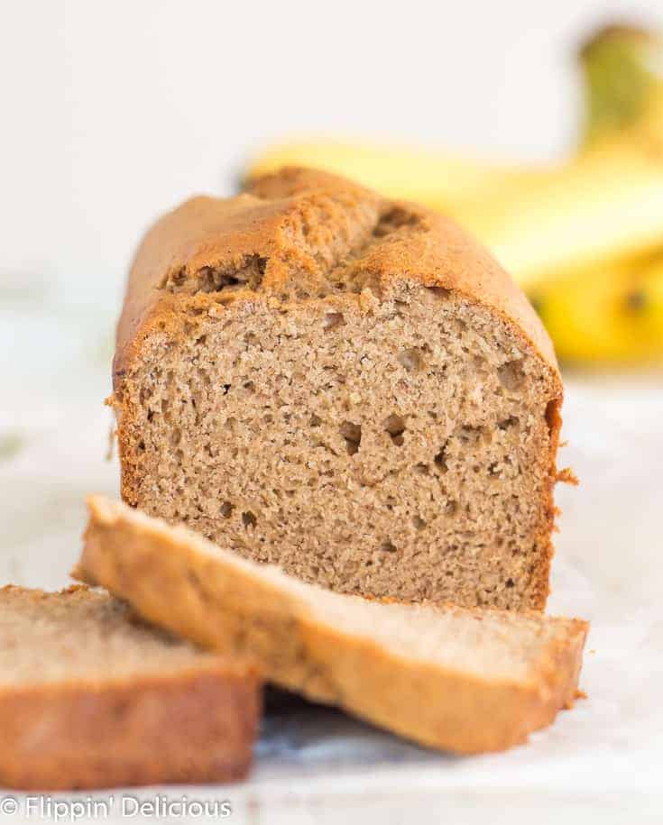 loaf of gluten free banana bread with a few slices in front and out of focus with a bunch of bananas in the background, on a white wooden table