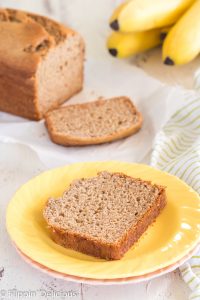 slice of gluten free banana bread on a yellow plate on top with a slice and full loaf of gluten free banana bread and bunch of ripe bananas in the background