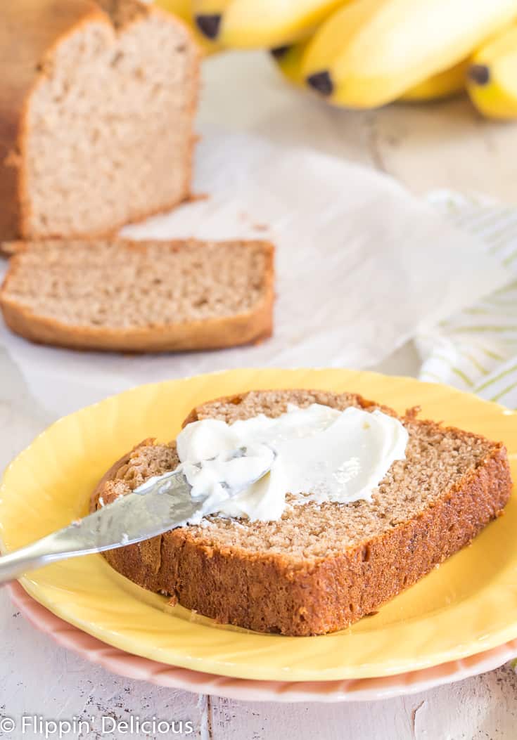 slice of gluten free banana bread on a yellow plate, small knife spreading cream cheese on top with a slice and full loaf of gluten free banana bread and bunch of ripe bananas in the background