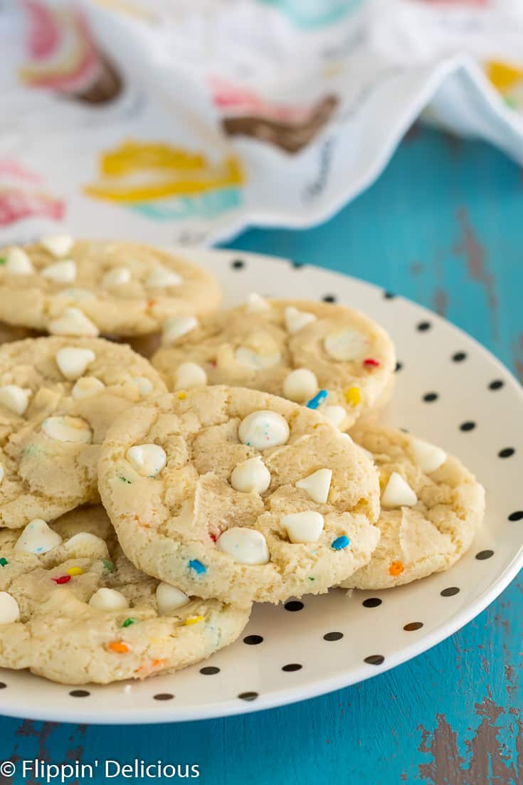 gluten free cookies with white chocolate chips and sprinkles on a cream plate with polka dots on top of a turquoise background with a cupcake dishtowel in the background