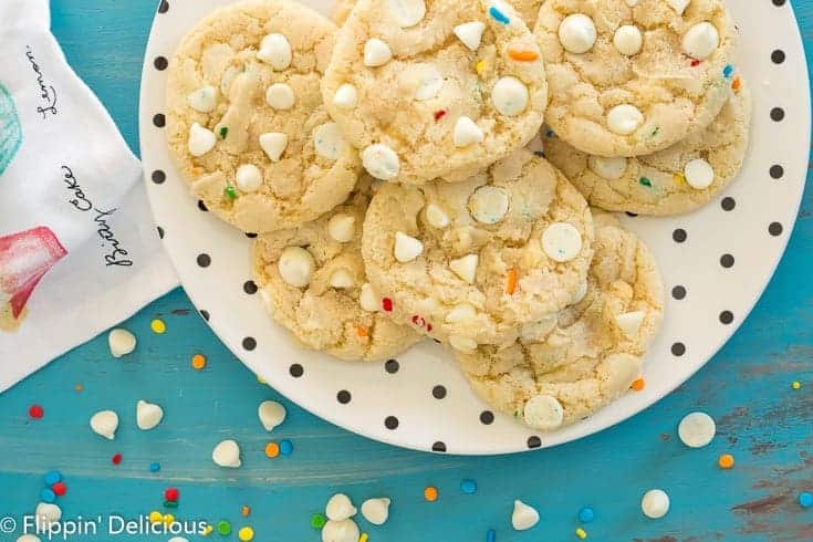 gluten free white chocolate chip cookies with sprinkles on a cream plate with black polka dots on a teal wooden table with white chocolate chips and rainbow sprinkles tossed on it