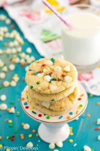 gluten free funfetti cookies with pudding mix on pedestal with white chocolate chips, sprinkles, and a glass of milk in the background