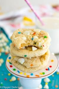 four gluten free funfetti cookies with white chocolate chips, with a bite taken out of the top cookie, on a pedestal on a teal background with a glass of milk, white chocolate chips, more sprinkles, and a dishtowel in the background