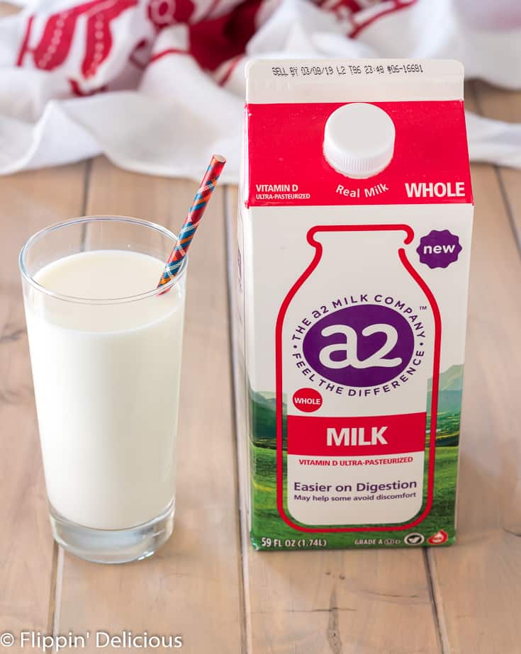 A2 milk in a glass beside carton of A2 milk on a wooden table with a red and white dish towel in the back