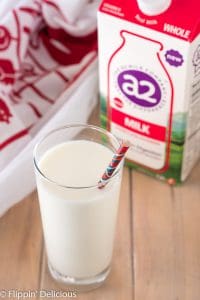 A2 milk in a glass in front of carton of A2 milk on a wooden table with a red and white dish towel in the back