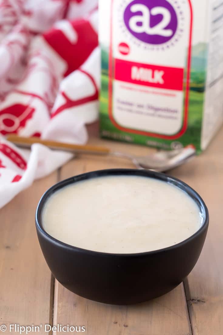 gluten free vanilla pudding in a black bowl on a wooden table with a carton of A2 milk and a red and white dish towel in the background