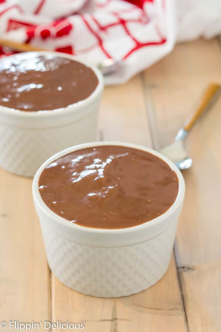 gluten free chocolate pudding in a ramekin with spoon, another ramekin of chocolate pudding, and a red and white dish towel in the background, all on a wooden table.