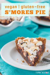 a slice of vegan gluten free s'mores pie with toasted marshmallows on a white plate with a fork, on a teal dish towel with a full pie and remaining slice in the background. with text "vegan | gluten-free s'mores pie"