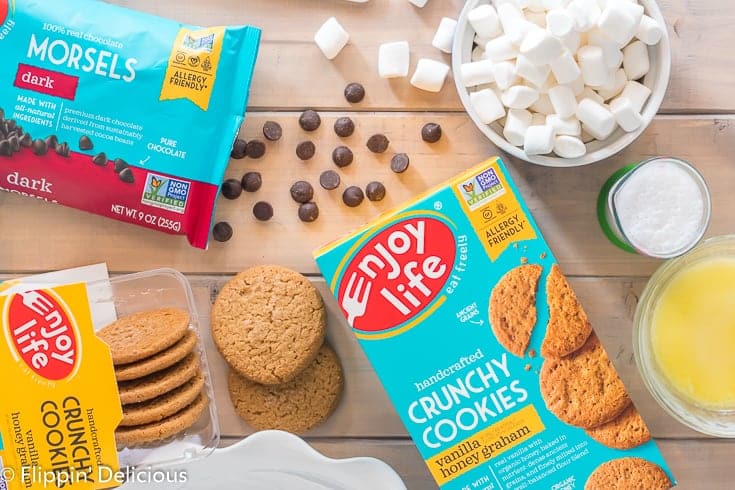 Overhead view of enjoy life vanilla honey graham crunchy cookies, dark chocolate morsels, vegan marshmallows, melted vegan butter, and dairy free milk on a wooden table