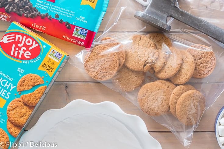 empty white pie plate with a bag full of crunchy vanilla honey graham cookies and a mallet with a box of enjoy life vanilla honey graham crunchy cookies and dark chocolate morsels