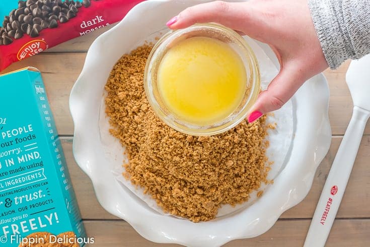 a hand holding a small glass bowl with melted vegan butter over a white ceramic pie plate with fluted edges, lined with a circle of parchment paper, filled with crushed enjoy life vanilla honey graham crunchy cookies with a box of cookies and bag of dark morsels on the wooden table