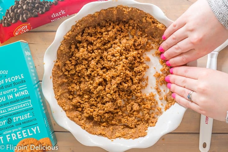 hands with pink painted finger nails pressing a gluten free graham cracker crust up the sides of a white ceramic pie plate with a box of cookies and bag of dark morsels on the wooden table