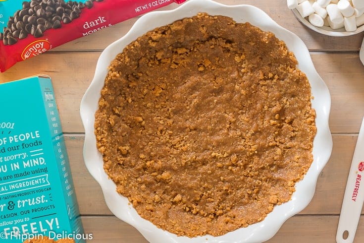 Finished no bake gluten free graham cracker pie crust on a wooden table with a box of enjoy life vanilla honey graham crunchy cookies and bag of dark chocolate morsels