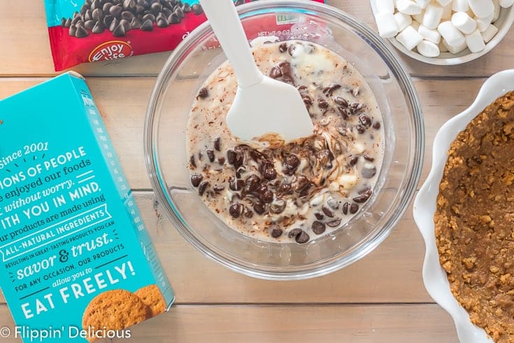 partially melted enjoy life dark morsels, vegan butter, and dairy free milk in a glass bowl with a white rubber spatula on a wooden table with enjoy life crunchy cookies and dark morsels in packaging on wooden table 