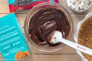 vegan fudge pie filling made with enjoy life dark morsels, vegan butter, and dairy free milk in a glass bowl with a white rubber spatula on a wooden table with enjoy life crunchy cookies and dark morsels in packaging