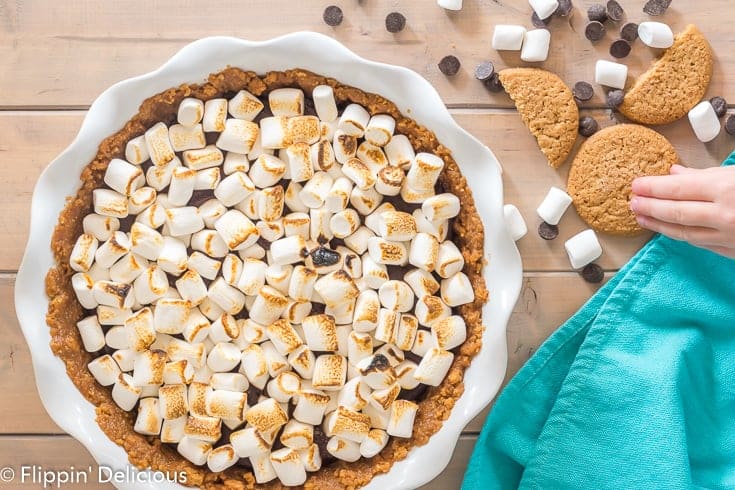 overhead vegan gluten free s'mores pie with toasted marshmallows on a wooden table with a teal dish towel and some cookies , marshmallows, and dark morsels