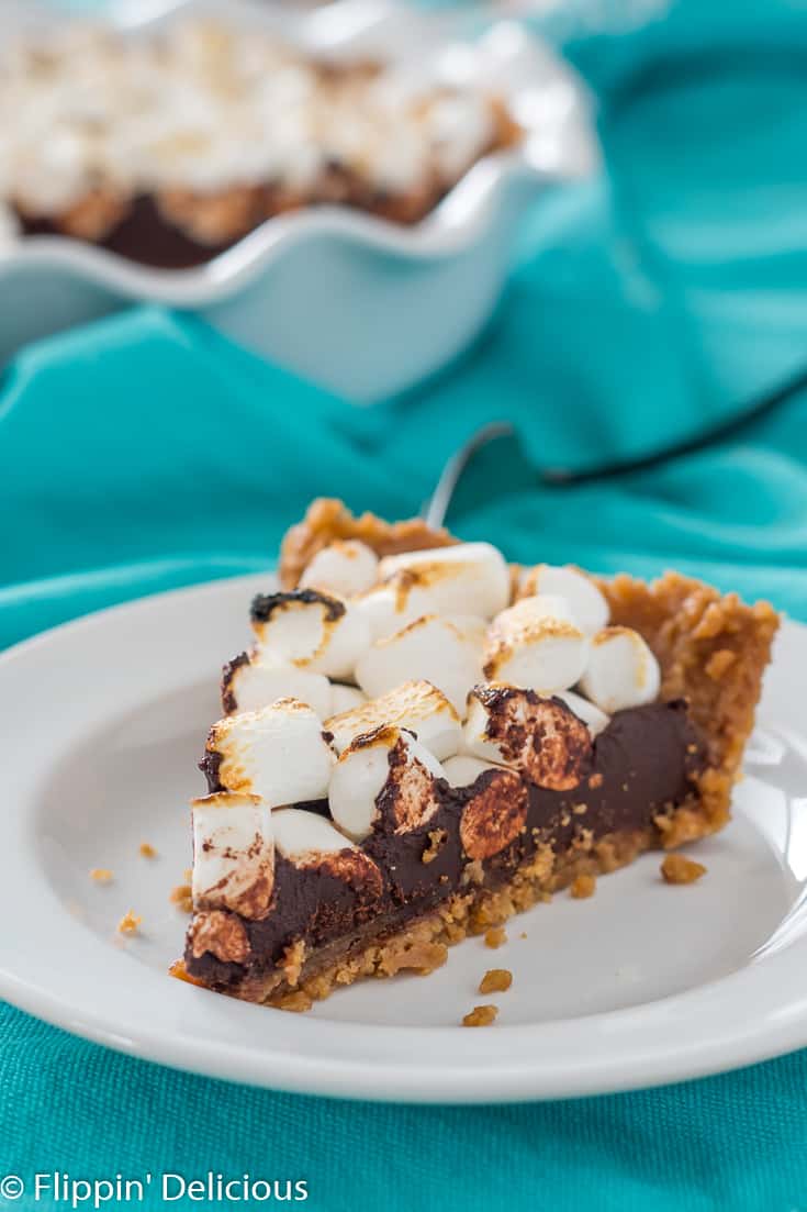 slice of vegan gluten free s'mores fudge pie with toasted marshmallows on a white plate with a teal dish towel and full pie in the background