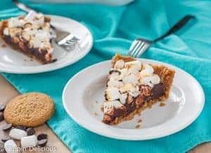 two slices of gluten free s'mores pie on white plates with a teal dish towel and some cookies, dark morsels, and vegan marshmallows