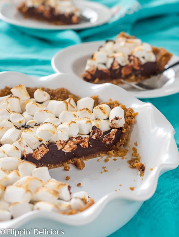 vegan gluten free s'mores pie with toasted marshmallows in a white ceramic pie dish with fluted edges, with another slice of pie on a plate with a fork, on a teal dish towel in the background.
