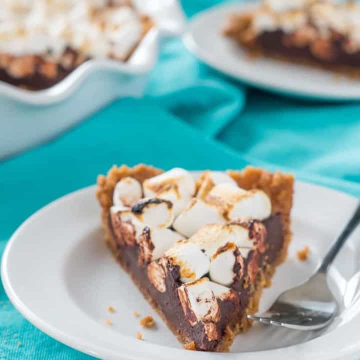 a slice of vegan gluten free s'mores pie with toasted marshmallows on a white plate with a fork, on a teal dish towel with a full pie and remaining slice in the background.