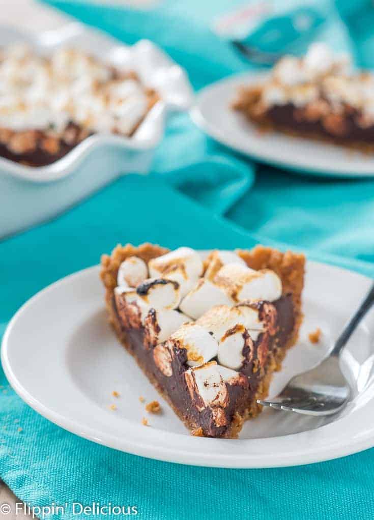 a slice of vegan gluten free s'mores pie with toasted marshmallows on a white plate with a fork, on a teal dish towel with a full pie and remaining slice in the background.