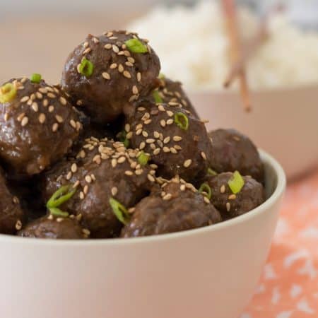 close up of gluten free teriyaki meatballs in a bowl garnished with sesame seeds and green onions with a blurry bowl of rice with chopsticks in the background.