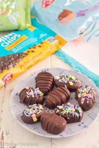 chocolate covered cookie dough truffles shaped like Easter eggs on a blue plate (some are drizzle with chocolate and some decorated with easter sprinkles) with an easter tea towel and a bag of enjoy life mini chips